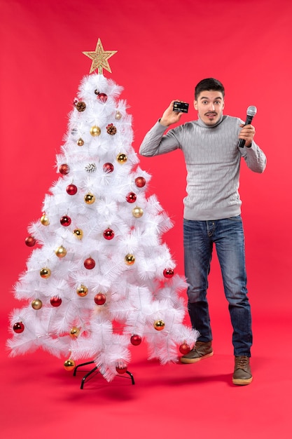 umore di Natale con ragazzo fiducioso vestito in jeans in piedi vicino all'albero di Natale decorato e tenendo il microfono e il suo telefono