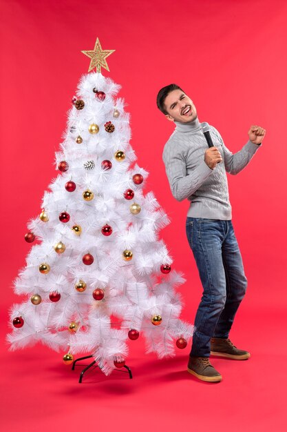 umore di Natale con ragazzo fiducioso vestito in jeans in piedi vicino all'albero di Natale decorato e tenendo il microfono