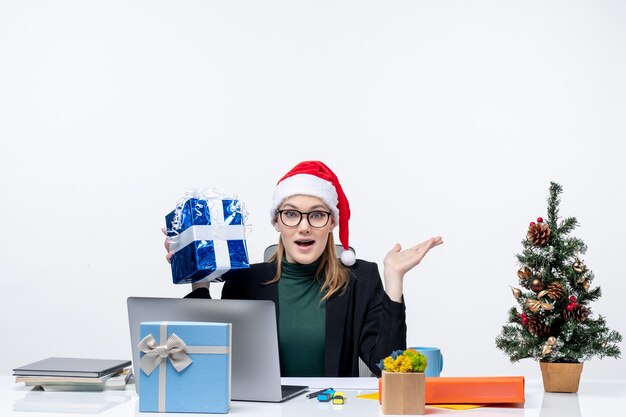 Umore di natale con sorprese giovane donna con cappello di babbo natale e occhiali da vista seduto a un tavolo tenendo il suo regalo su sfondo bianco