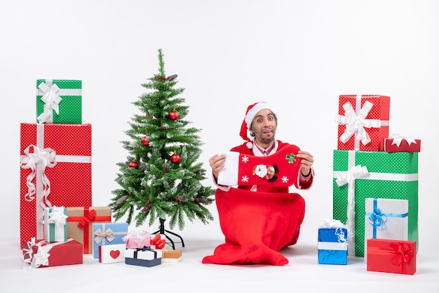Umore di nuovo anno con Babbo Natale positivo divertente seduto per terra e tenendo il calzino di Natale vicino a regali e albero di Natale decorato su sfondo bianco