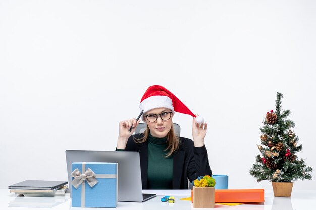 Umore di nuovo anno con donna bionda confusa con un cappello di Babbo Natale seduto a un tavolo con un albero di Natale e un regalo su di esso su sfondo bianco
