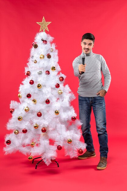 Umore di nuovo anno con ragazzo positivo vestito in jeans in piedi vicino all'albero di Natale decorato