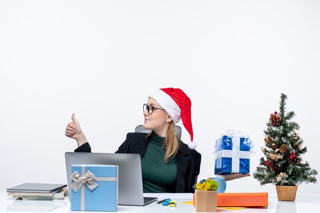 Umore festivo con la giovane donna sorpresa con il cappello di babbo natale e gli occhiali da portare seduti a un tavolo che mostra il regalo di natale che indica qualcosa sul lato destro che fa il gesto giusto