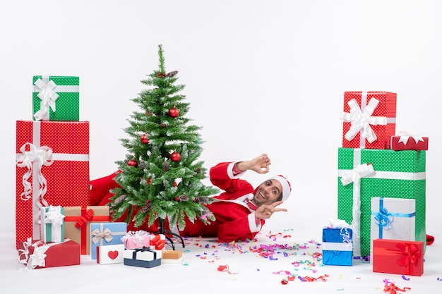 Umore festivo di festa con il giovane Babbo Natale che si trova dietro l'albero di Natale e che mostra due regali vicini su priorità bassa bianca