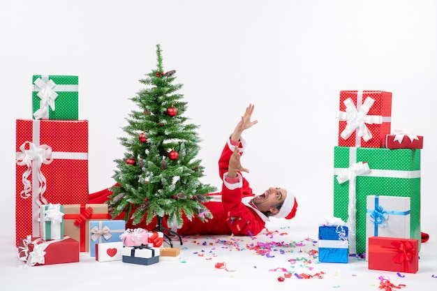 Umore festivo di festa con il giovane Babbo Natale emotivo che si trova dietro l'albero di Natale vicino a regali su priorità bassa bianca