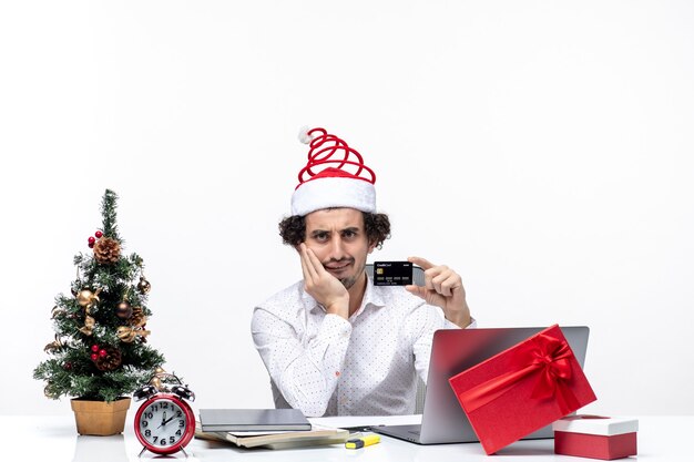 Umore festivo di vacanza con uomo d'affari confuso con cappello di Babbo Natale che tiene carta di credito e mette la mano sotto il mento in ufficio