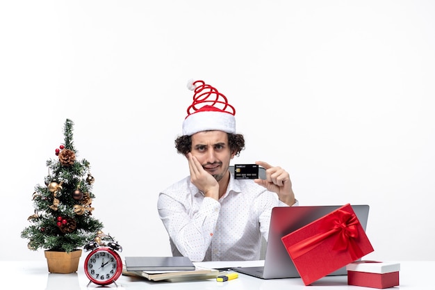 Umore festivo di vacanza con uomo d'affari confuso con cappello di Babbo Natale che tiene carta di credito e mette la mano sotto il mento in ufficio