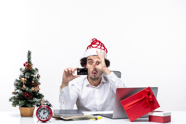 Umore festivo vacanza con giovane uomo d'affari divertente con cappello di Babbo Natale e tenendo la sua carta di credito in ufficio su priorità bassa bianca
