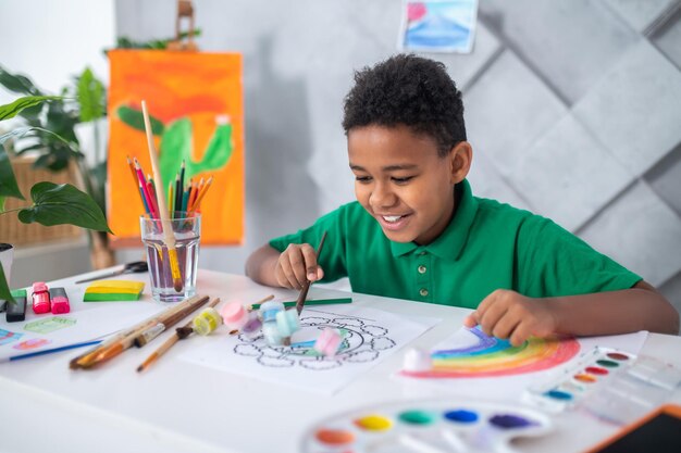 Umore giocoso. Felice ragazzo dalla pelle scura di età scolare in maglietta verde seduto a tavola con forniture d'arte che spingono tubi di vernice in piedi nella foto in una stanza luminosa