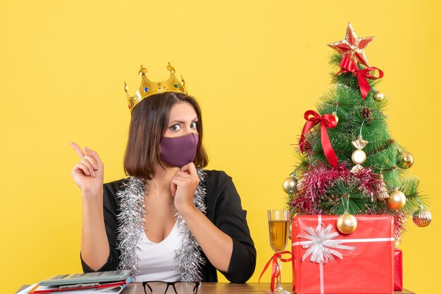 Umore natalizio con bella signora concentrata in vestito con corona che indossa la sua maschera medica in ufficio su giallo