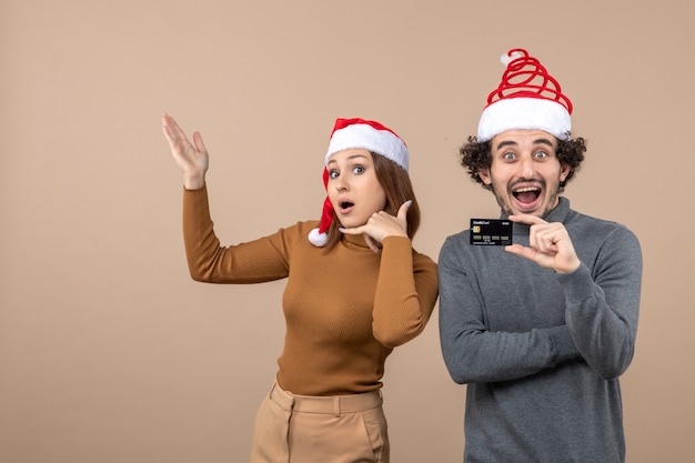 umore natalizio con coppia cool eccitata e soddisfatta che indossa cappelli rossi di Babbo Natale che mostra carta di credito e donna che fa gesto di chiamata