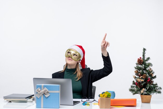 Umore natalizio con eccitata giovane donna positiva con cappello di Babbo Natale e maschera da portare seduto a un tavolo che punta sopra su uno sfondo bianco