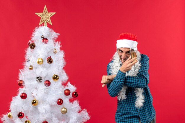 umore natalizio con felice giovane pazzo con cappello di Babbo Natale in una camicia blu spogliata, alzando un bicchiere di vino vicino all'albero di Natale