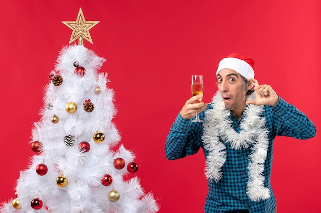umore natalizio con giovane emotivo con cappello di Babbo Natale in una camicia blu spogliata ascoltando attentamente tenendo un bicchiere di vino vicino all'albero di Natale