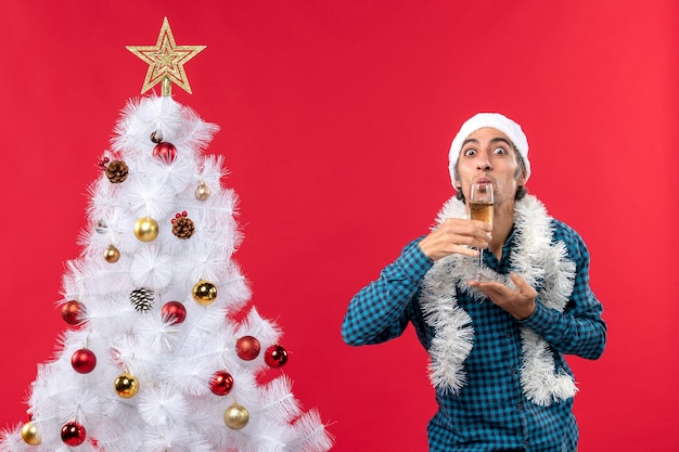 umore natalizio con giovane emotivo con cappello di Babbo Natale in una camicia blu spogliata che tiene e beve un bicchiere di vino vicino all'albero di Natale