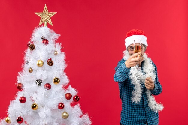 umore natalizio con giovane emotivo con cappello di Babbo Natale in una camicia blu spogliata che tiene e mostra un bicchiere di vino vicino all'albero di Natale