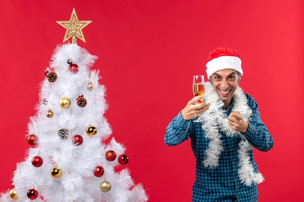 umore natalizio con giovane emotivo con cappello di Babbo Natale in una camicia blu spogliata che tiene un bicchiere di vino che punta qualcosa vicino all'albero di Natale