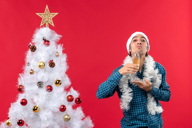 umore natalizio con giovane emotivo con cappello di babbo natale in una maglietta blu spogliata che tiene un bicchiere di vino sotto il mento vicino all'albero di Natale