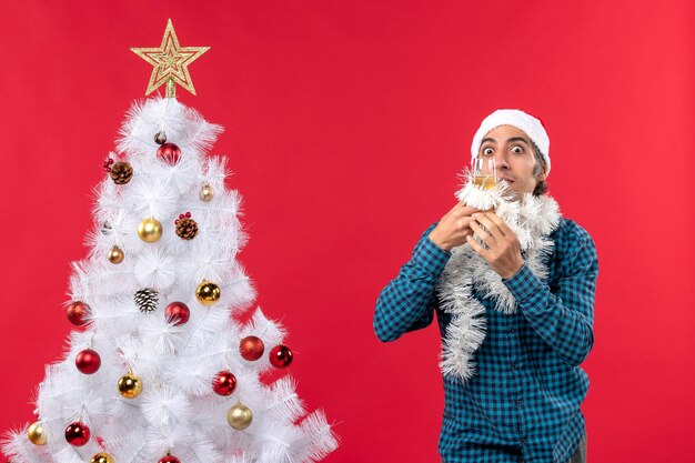 umore natalizio con giovane uomo con cappello di Babbo Natale e alzando un bicchiere di vino si rallegra vicino all'albero di Natale