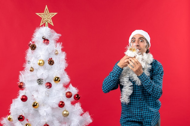 umore natalizio con giovane uomo con cappello di Babbo Natale e alzando un bicchiere di vino si rallegra vicino all'albero di Natale