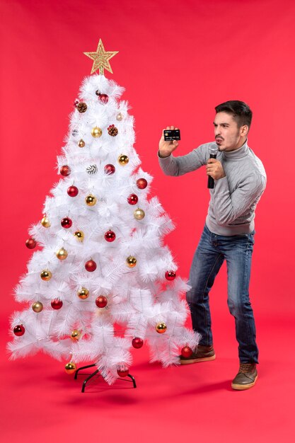 umore natalizio con ragazzo emotivo in piedi vicino all'albero di Natale decorato e tenendo il microfono e il telefono con gesti divertenti