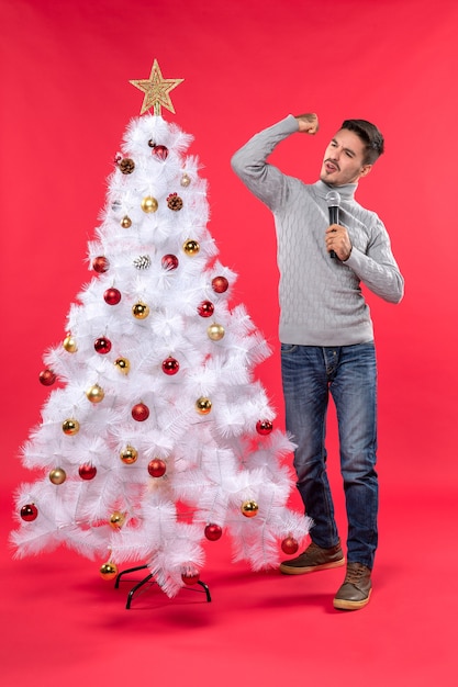 umore natalizio con ragazzo positivo vestito in jeans in piedi vicino all'albero di Natale decorato e tenendo il microfono che mostra il suo potere