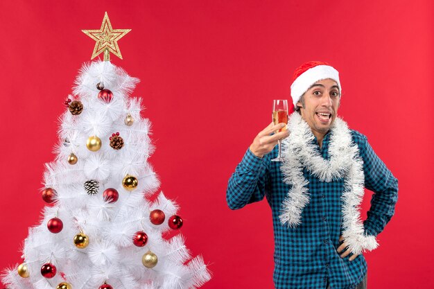 umore natalizio con un giovane emotivo con cappello di Babbo Natale e alzando un bicchiere di vino si rallegra vicino all'albero di Natale