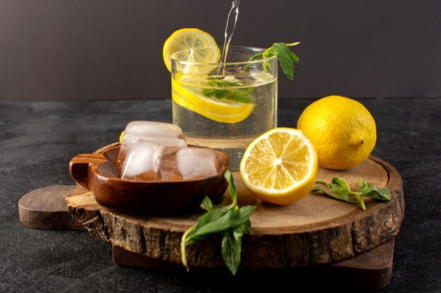 Un'acqua vista frontale con bevanda fresca fresca di limone all'interno di vetro versando con cubetti di ghiaccio con fette di limoni sul buio