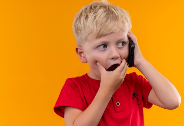 Un adorabile ragazzino con i capelli biondi e gli occhi azzurri che indossa la maglietta rossa che parla al telefono cellulare mentre guarda sorprendentemente lato con la mano sulla bocca