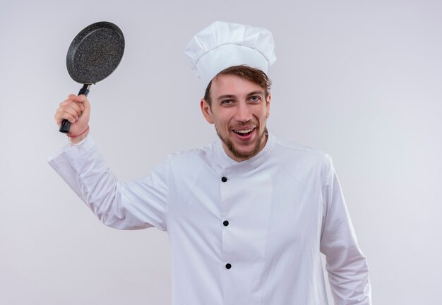 Un allegro chef biondo uomo che indossa l'uniforme bianca del fornello e il cappello che tiene la padella come una mazza da baseball su un muro bianco