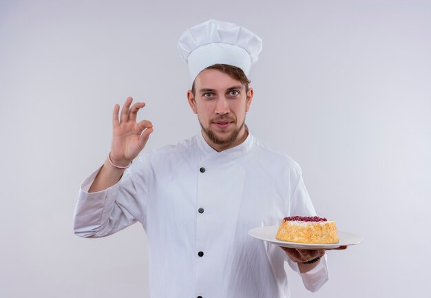 Un allegro giovane chef barbuto uomo che indossa l'uniforme bianca del fornello e il cappello che tiene un piatto con la torta e mostra un gustoso gesto ok mentre guarda un muro bianco