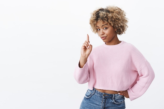 Un'altra cosa, ti insegno. Ritratto di intelligente e creativa bella donna africana sicura di sé con capelli biondi ricci sogghignando e alzando il dito indice dando indicazioni e saggi consigli