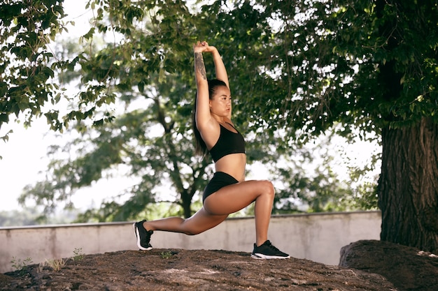 Un atleta femminile muscolare che fa allenamento al parco. Ginnastica, allenamento, flessibilità allenamento fitness. Città estiva in una giornata di sole sul campo di sfondo. Stile di vita attivo e sano, gioventù, bodybuilding.