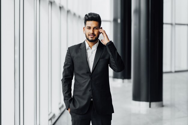 Un attraente uomo d'affari indiano barbuto in un moderno centro di uffici, che indossa un abito nero, tiene la mano vicino a un auricolare wireless