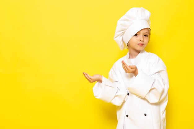 Un bambino sveglio di vista frontale in vestito bianco del cuoco e cappuccio bianco del cuoco che posano sull'alimento giallo della cucina del cuoco del bambino della parete gialla