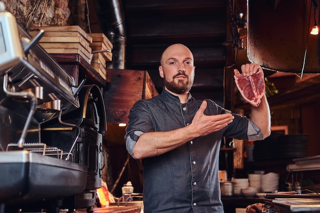 Un bel chef barbuto che presenta una bistecca fresca prima di cucinare e guarda una telecamera nella cucina di un ristorante.