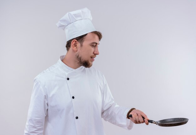 Un bel giovane chef barbuto uomo che indossa l'uniforme bianca fornello e cappello guardando la padella su un muro bianco