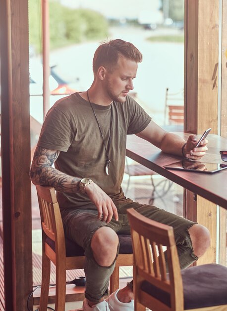 Un bel hipster alla moda si siede a un tavolo in un caffè lungo la strada, leggendo il messaggio sullo smartphone.