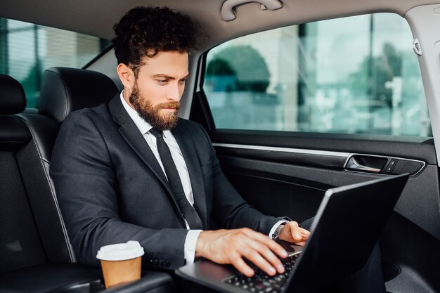 Un bel manager barbuto che lavora sul suo laptop con il caffè per andare sul sedile posteriore della nuova auto