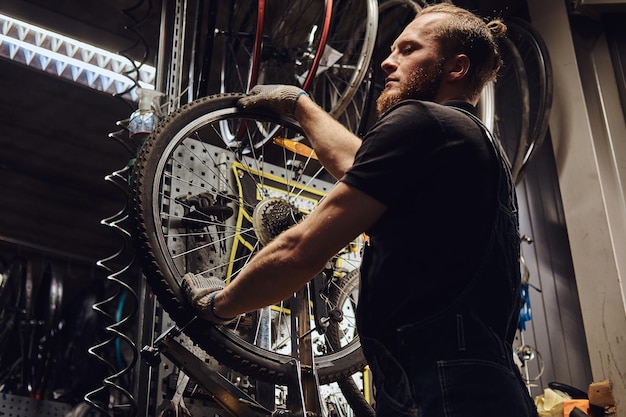 Un bel maschio rosso con una tuta di jeans, che lavora con una ruota di bicicletta in un'officina. Un lavoratore rimuove il pneumatico della bicicletta in un'officina.