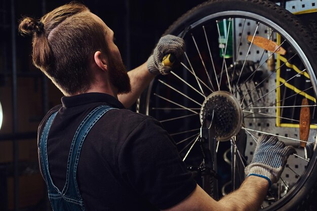 Un bel maschio rosso con una tuta di jeans, che lavora con una ruota di bicicletta in un'officina. Un lavoratore rimuove il pneumatico della bicicletta in un'officina.