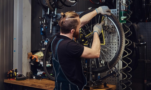 Un bel maschio rosso con una tuta di jeans, che lavora con una ruota di bicicletta in un'officina. Un lavoratore rimuove il pneumatico della bicicletta in un'officina.