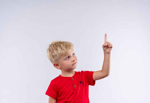 Un bel ragazzino con i capelli biondi e gli occhi azzurri che indossa la maglietta rossa rivolta verso l'alto con il dito indice su una parete bianca