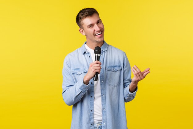 Un bel ragazzo biondo sfacciato in abiti casual esegue discorsi, spettacolo in piedi davanti al pubblico, cantando una canzone e sorridente impertinente, in piedi sfondo giallo, godendosi la notte di karaoke, tenendo il microfono.