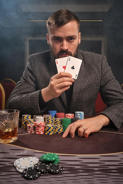 Un bel ragazzo con la barba in un abito classico sta giocando a poker seduto al tavolo del casinò in fumo. Sta mostrando due assi alla telecamera e si rende conto che ha vinto. Gioco d'azzardo per soldi. Giochi di per