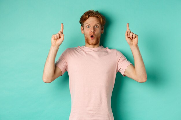 Un bel ragazzo impressionato con i capelli rossi che punta le dita in alto, dimostra l'offerta promozionale, in piedi in maglietta su sfondo di menta