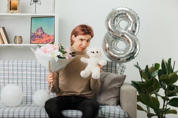 Un bel ragazzo impressionato durante la giornata delle donne felici che tiene in mano un bouquet con un orsacchiotto seduto sul divano in soggiorno