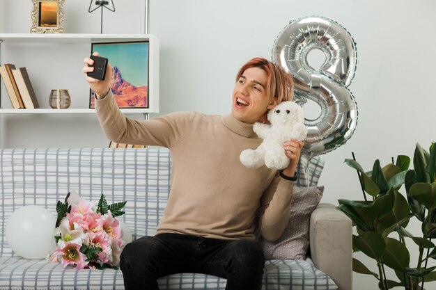 Un bel ragazzo sorridente durante la giornata delle donne felici che tiene l'orsacchiotto si fa un selfie seduto sul divano in soggiorno