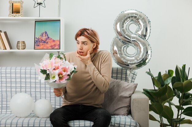 Un bel ragazzo spaventato ha afferrato il mento durante la giornata delle donne felici che tiene e guarda il bouquet seduto sul divano nel soggiorno