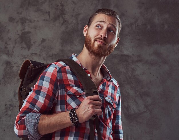 Un bel viaggiatore rosso alla moda in una camicia di flanella con uno zaino, in posa in uno studio su sfondo grigio.